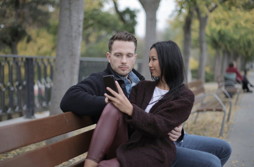 Positive young multiethnic couple using smartphone while sitting on bench in park