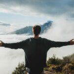 Back view of anonymous male traveler in casual clothes standing on edge of cliff and admiring breathtaking scenery of clouds and mountain top